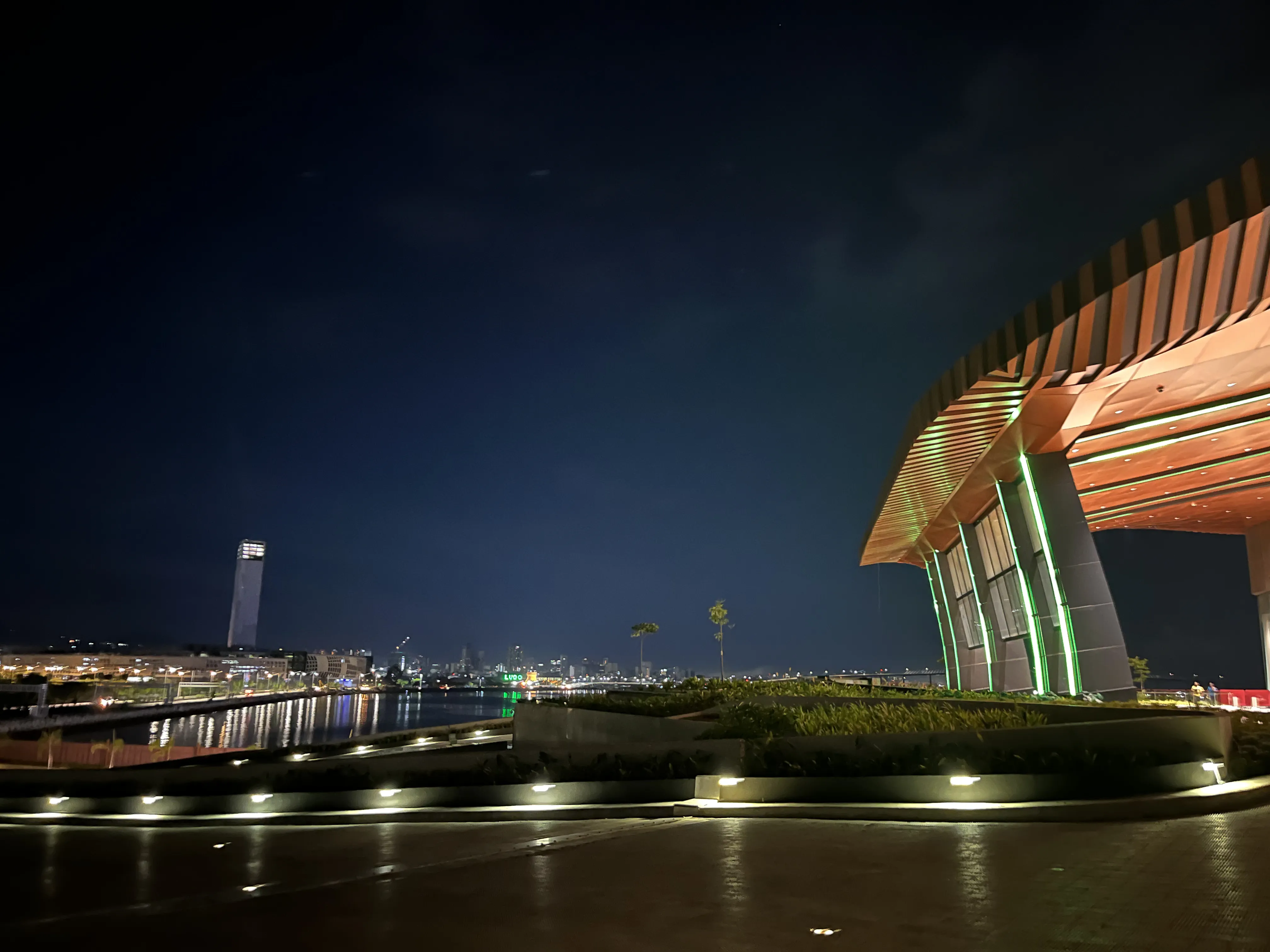 Modern convention venue on Manila Bay at night, near the site of ASEAN Tech Summit 2026 discussions Night view of a modern waterfront convention venue with a curved, wood-slat roof and green vertical accent lighting, overlooking Manila Bay with the city skyline glowing in the distance
